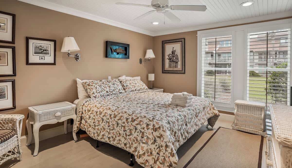A bedroom with a bed, nightstands, wall art, and a view through large windows.