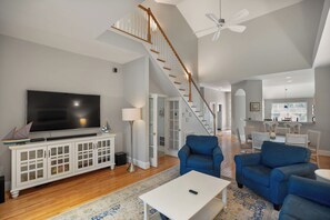A spacious living room with a large flat-screen TV, a white entertainment unit, blue armchairs, and a staircase leading to an upper level. - Stunning location ON Ocean Creek Golf Course! (Fripp Island)