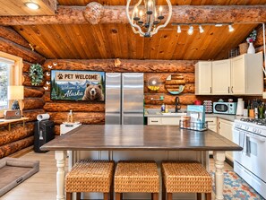 A log cabin interior with a central dining table, wooden stools, a refrigerator, a microwave, and a stove.