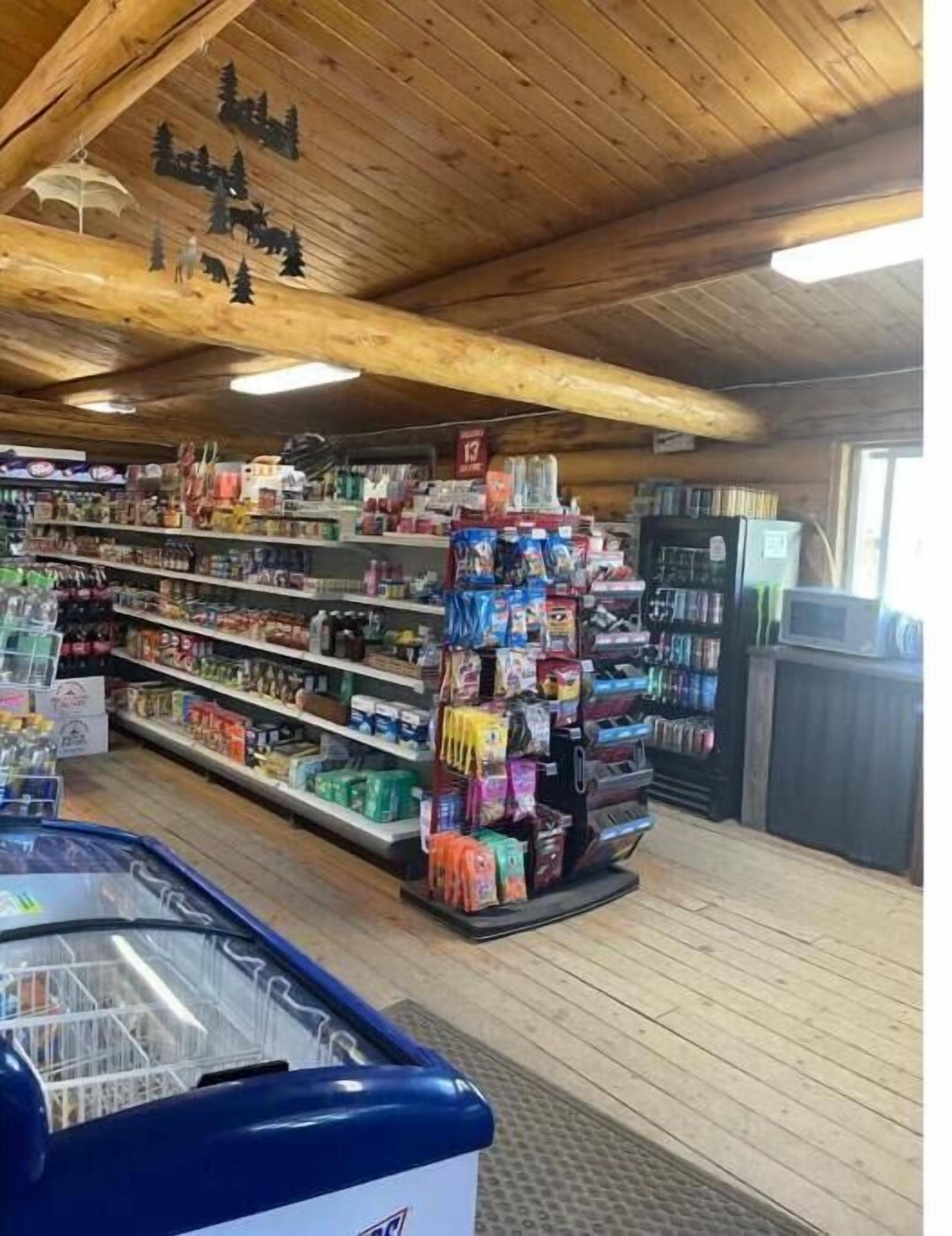 Una tienda con vigas de madera en el techo, estantes llenos de diversos productos y un mostrador refrigerado.
