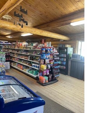 Una tienda con vigas de madera en el techo, estantes llenos de diversos productos y un mostrador refrigerado.