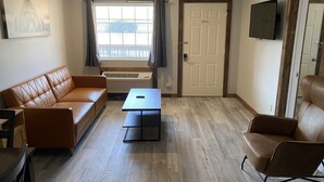 A living room with a brown leather sofa, a small table, a brown armchair, a window with curtains, a door, and a mounted TV. - 2BD 2BA w/Kitchenette close to Silver Dollar City (Branson)