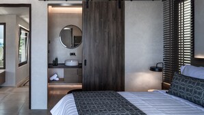 Um quarto moderno com uma cama grande, uma porta deslizante de madeira escura, um espelho redondo, uma mesa de cabeceira e uma janela com persianas.