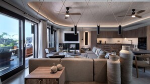 A modern living room with a large sofa, a dining table, and a kitchen area with wooden cabinets and a flat-screen TV.