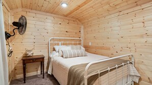 Un dormitorio con paneles de madera, una cama, una mesita de noche y un ventilador fijado a la pared.