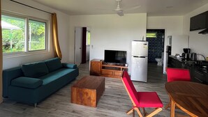 A modern living room with a sofa, coffee table, and red chairs. A kitchen is visible in the background with a refrigerator and stove. - Ânani - Mirimiri Lodge (Raiatea)