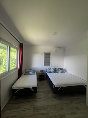 A small, well-lit room with two single beds, a window with curtains, a bedside table with a lamp, and an air conditioning unit on the wall. - Ânani - Mirimiri Lodge (Raiatea)
