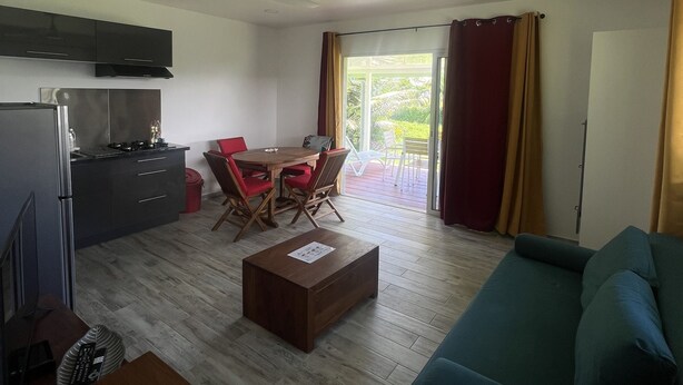 A modern living room with a sofa, a dining table with chairs, a kitchen area with cabinets and a refrigerator, and a sliding glass door leading to an outdoor area. - Ânani - Mirimiri Lodge (Raiatea)