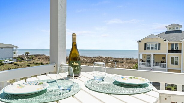Un balcón con una mesa para dos, con vista a la playa y una casa.