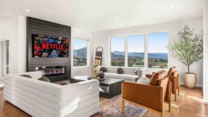 Ein modernes Wohnzimmer mit Kamin, einem großen Fernseher, der Netflix zeigt, und Bergblick durch große Fenster.