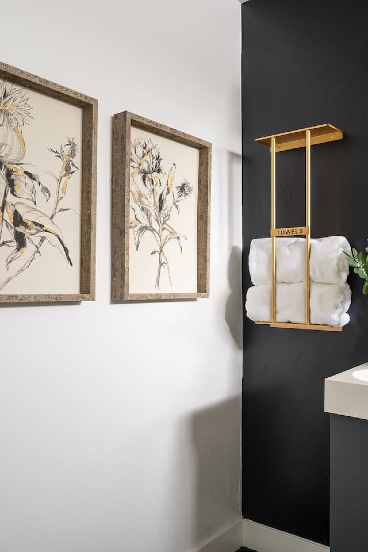 A bathroom with two framed botanical prints, a towel rack with folded towels, and a white sink.