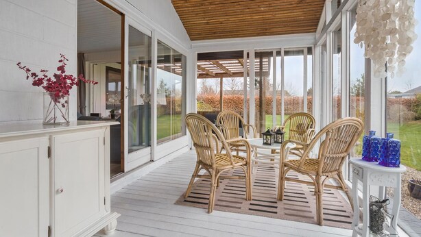 A covered patio with wicker chairs and a table, white walls, and a view of the garden.