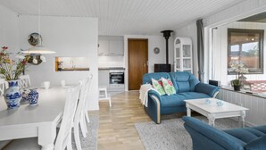 A modern living room with a blue sofa, white dining table, and chairs. There is a kitchen area with white cabinets and a stove. A large window provides natural light.