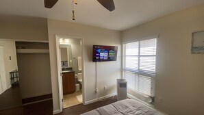 Un dormitorio con una cama, un televisor montado en la pared, un ventilador de techo y una ventana con persianas.
