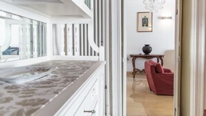 Una habitación con un ventanal grande, una silla roja, una pequeña mesa con un florero y una araña.