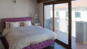 Un dormitorio con un cabecero tapizado color morado, una colcha blanca y un amplio ventanal corredera que da a un balcón.