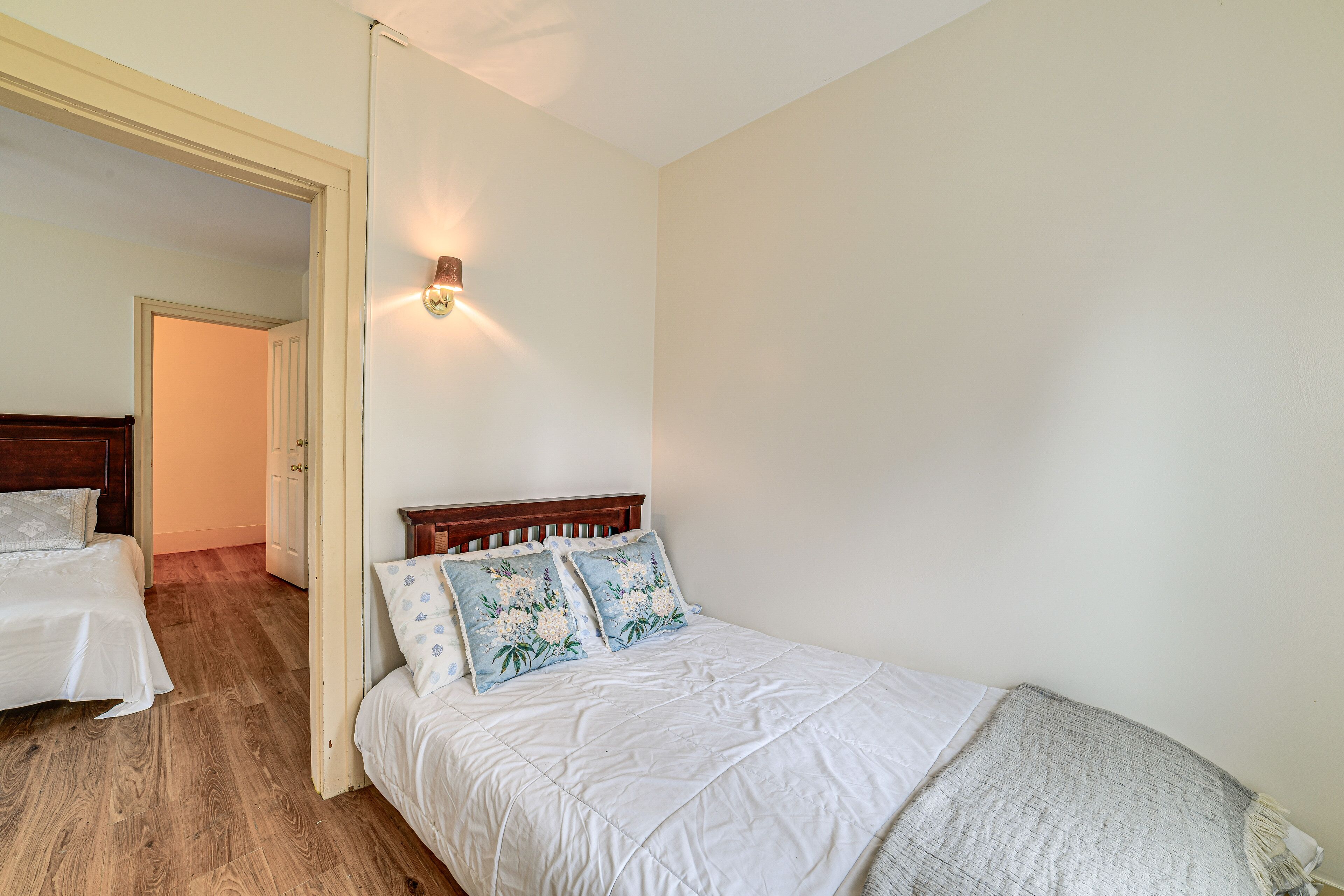 A bedroom with a wooden floor, a white bed with floral pillows, and a nightstand.