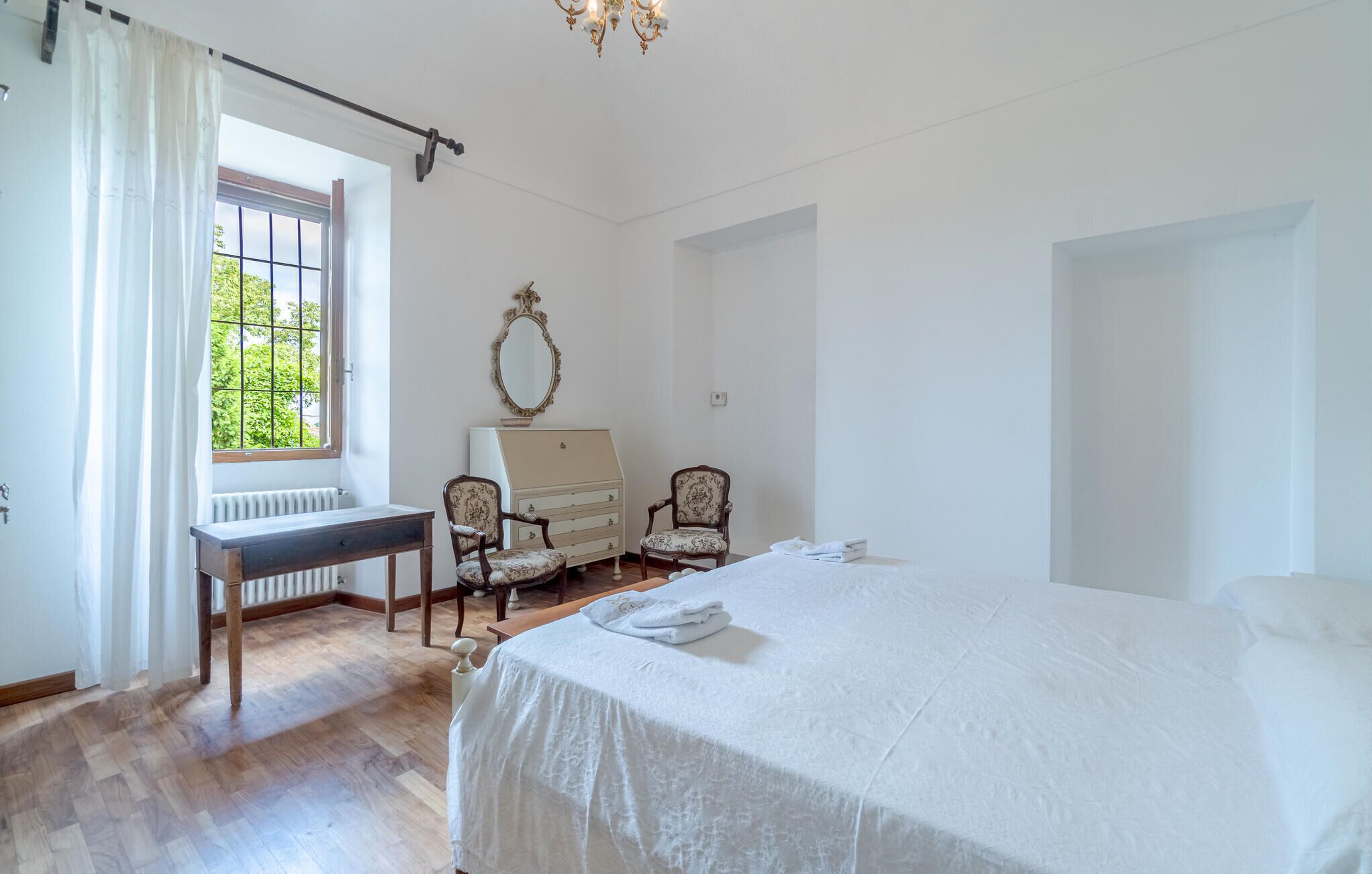 A bedroom with a large bed, a desk, two chairs, a dresser, and a window with curtains.