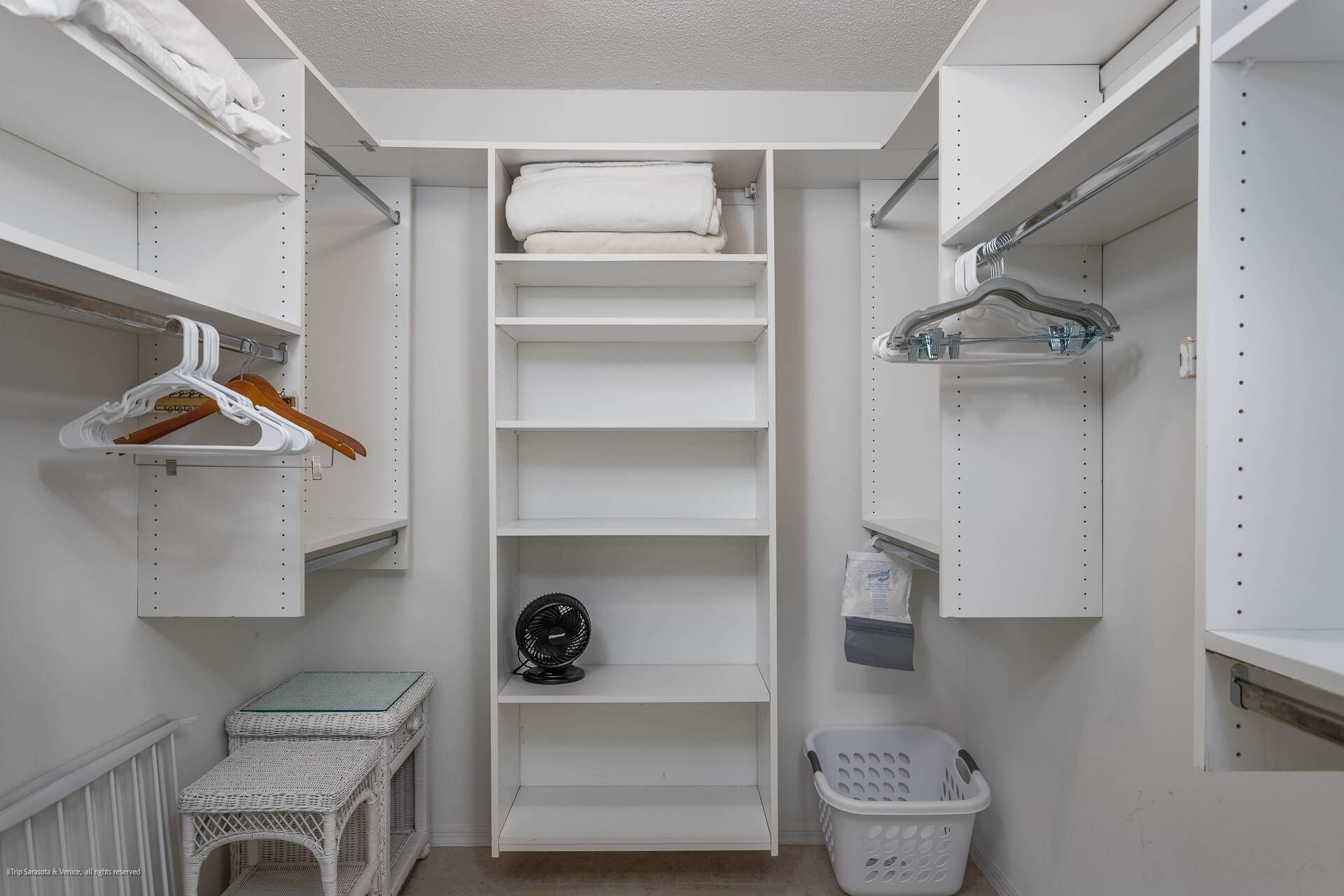 A walk-in closet with white shelves, a fan, a laundry basket, and a wicker table.