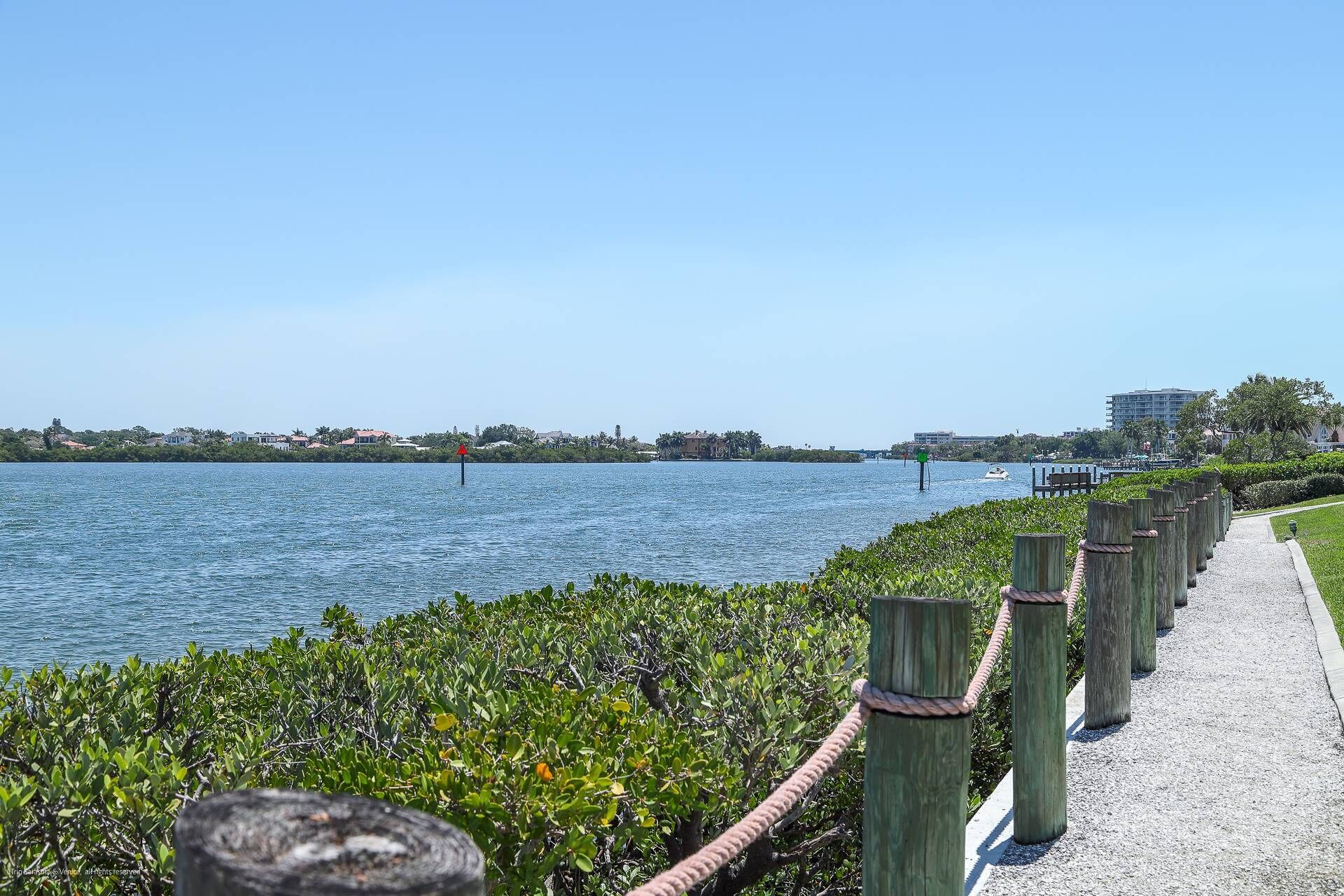 A waterfront promenade with a wooden fence, a grassy path, and a view of the water and distant buildings.