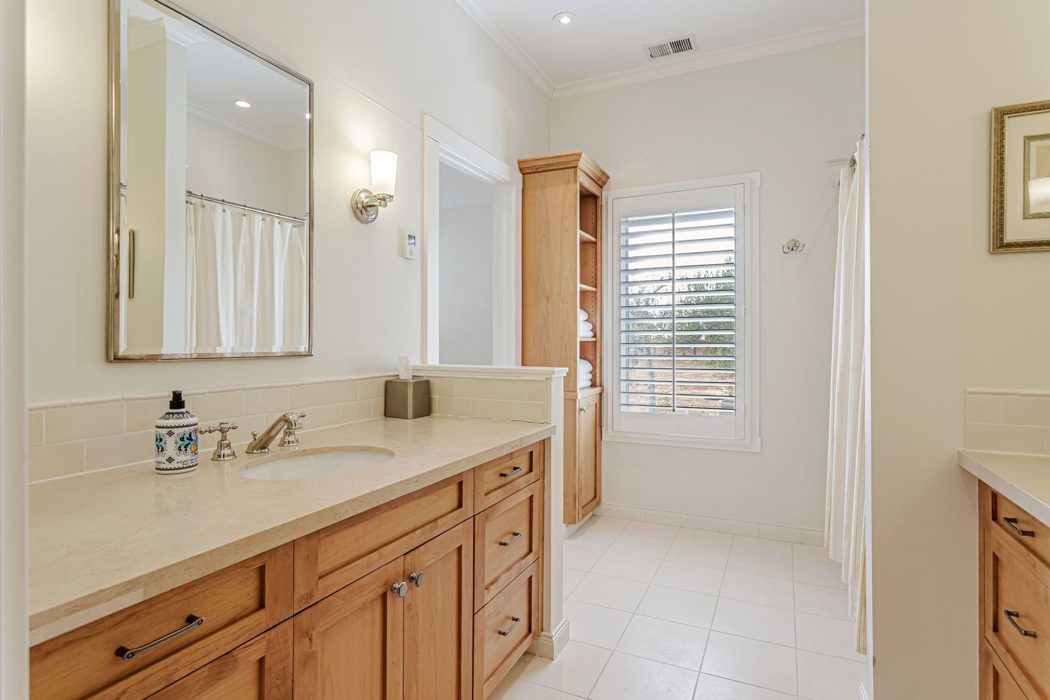 A bathroom with a large mirror, two sinks, wooden cabinets, and a window with blinds.