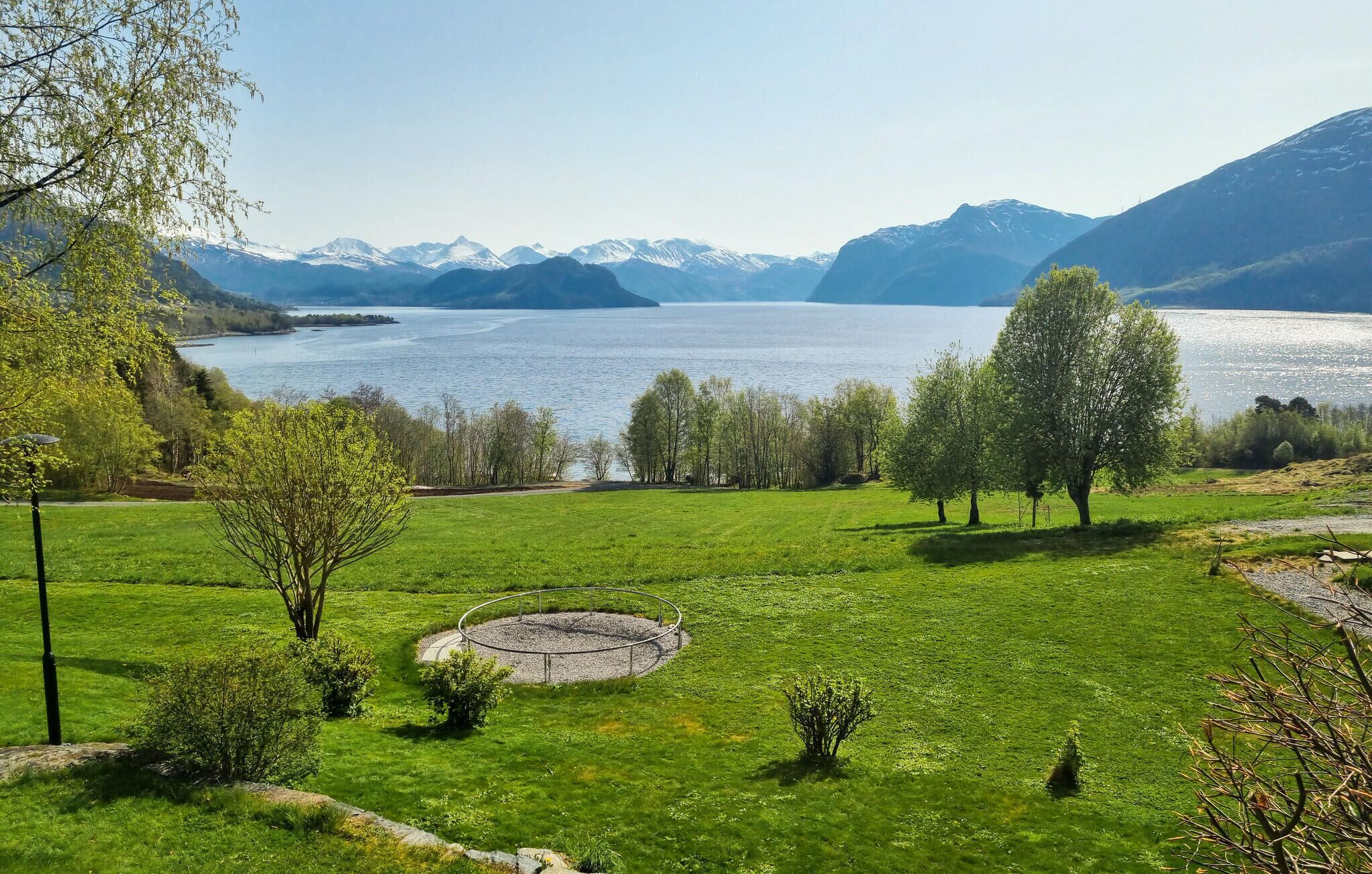 A serene lakeside landscape with a circular stone structure in the foreground, surrounded by lush green grass and trees.