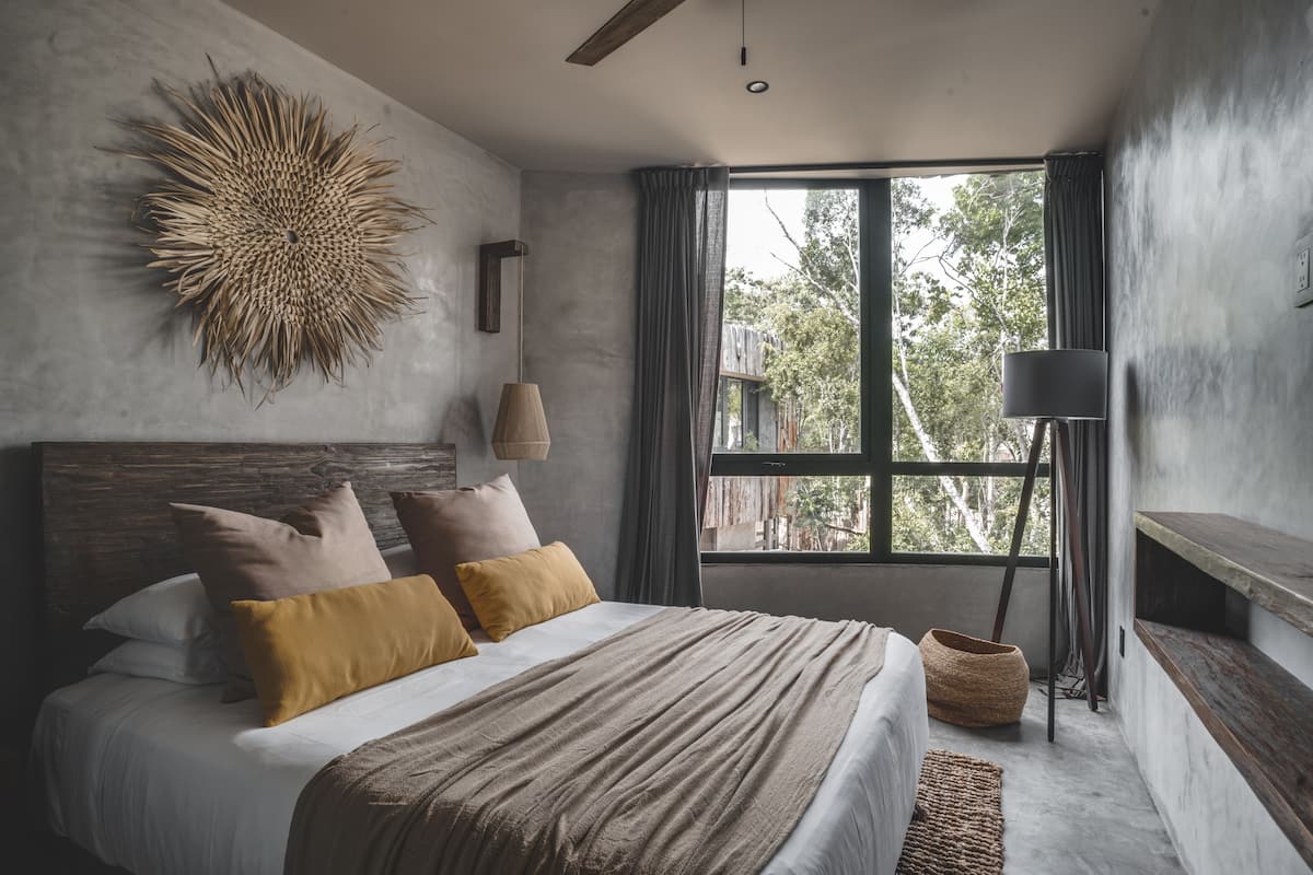 Quarto moderno com uma cama grande, uma obra de arte em tear na parede, um abajur e vista para árvores pela janela.