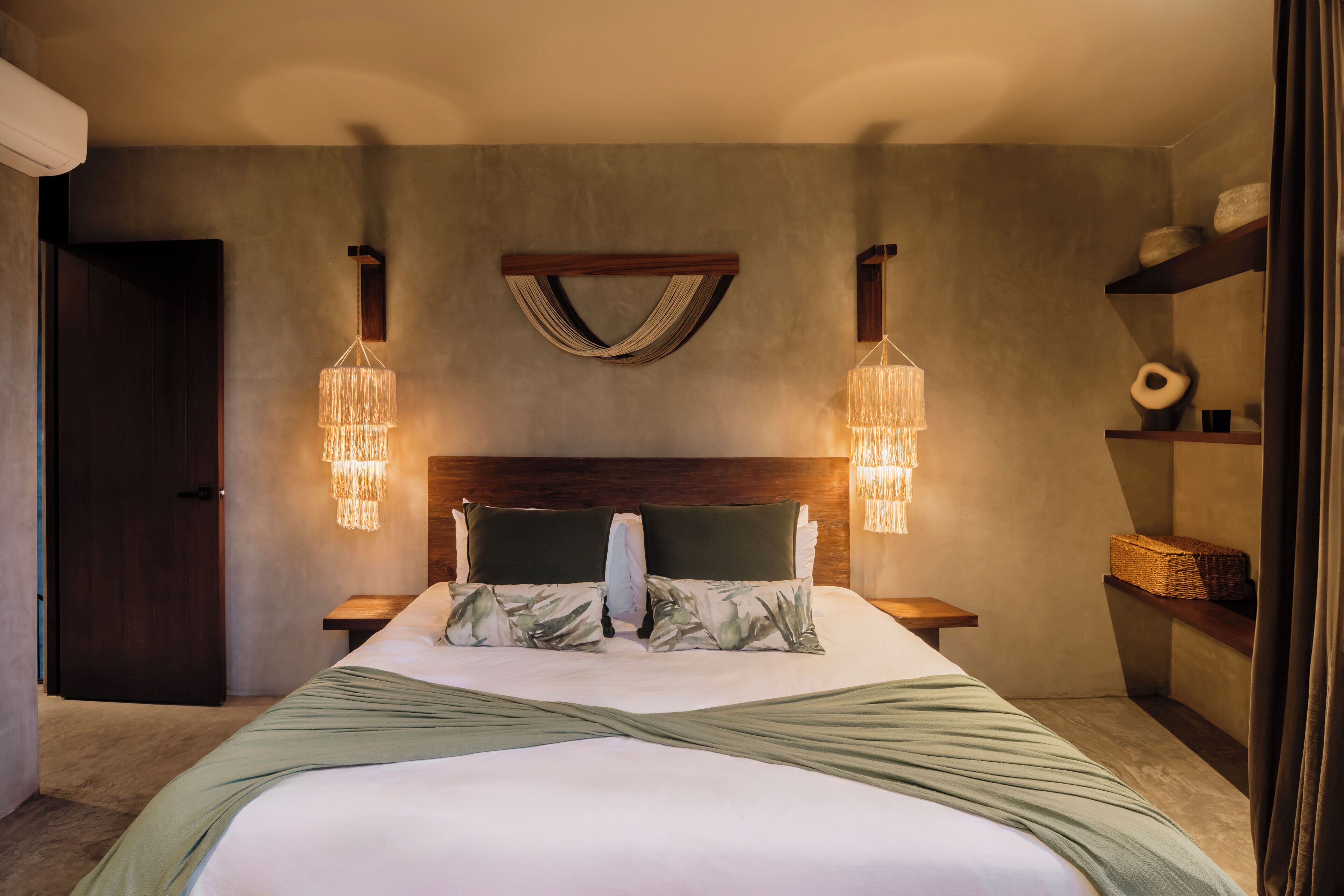 A bedroom with a large bed, two bedside tables, a wooden headboard, a wall-mounted light fixture, and a shelf with decorative items.