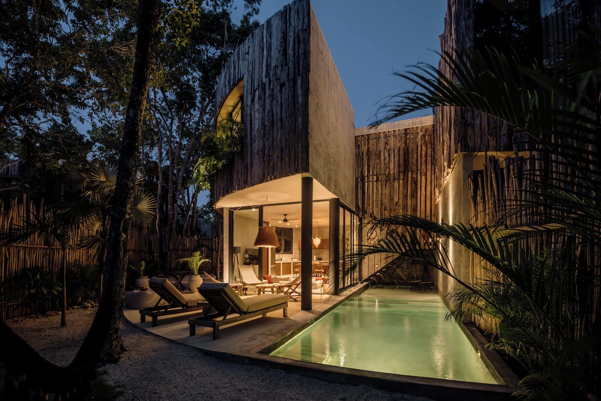 Área de piscina moderna ao ar livre, com espreguiçadeiras e um espaço de estar coberto, rodeada por plantas tropicais e um cercado de madeira.