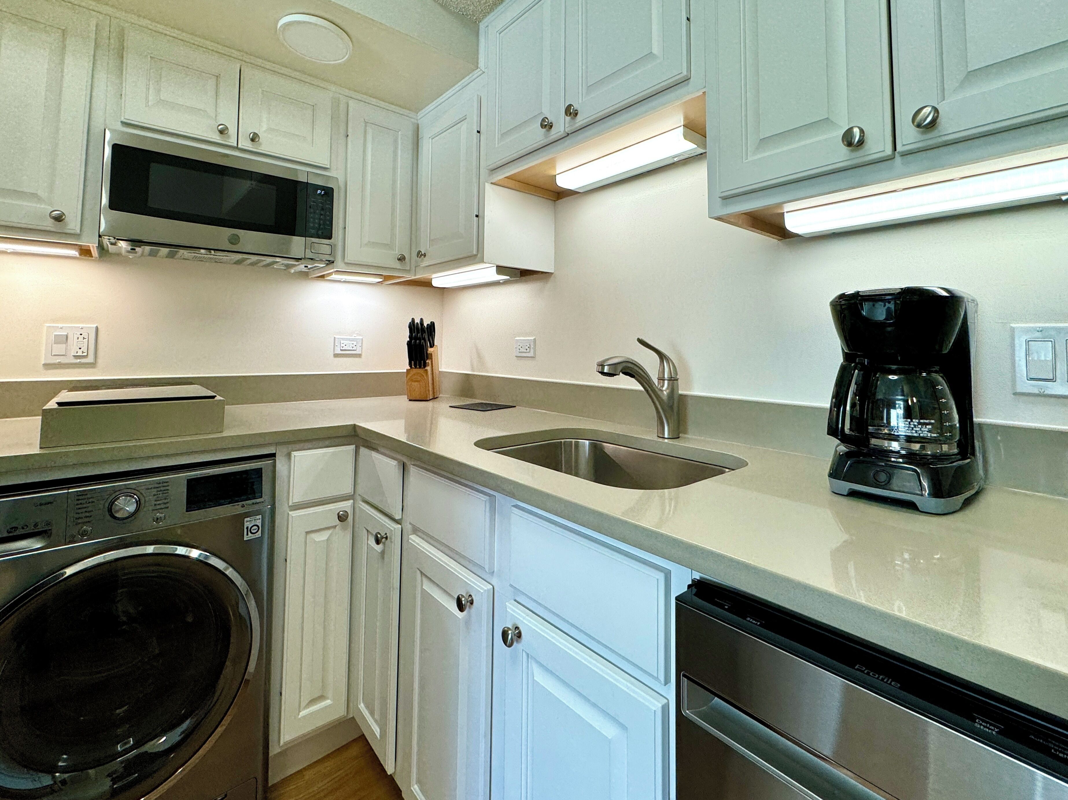 A compact kitchen with white cabinets, a stainless steel sink, a coffee maker, a microwave, and a dishwasher.