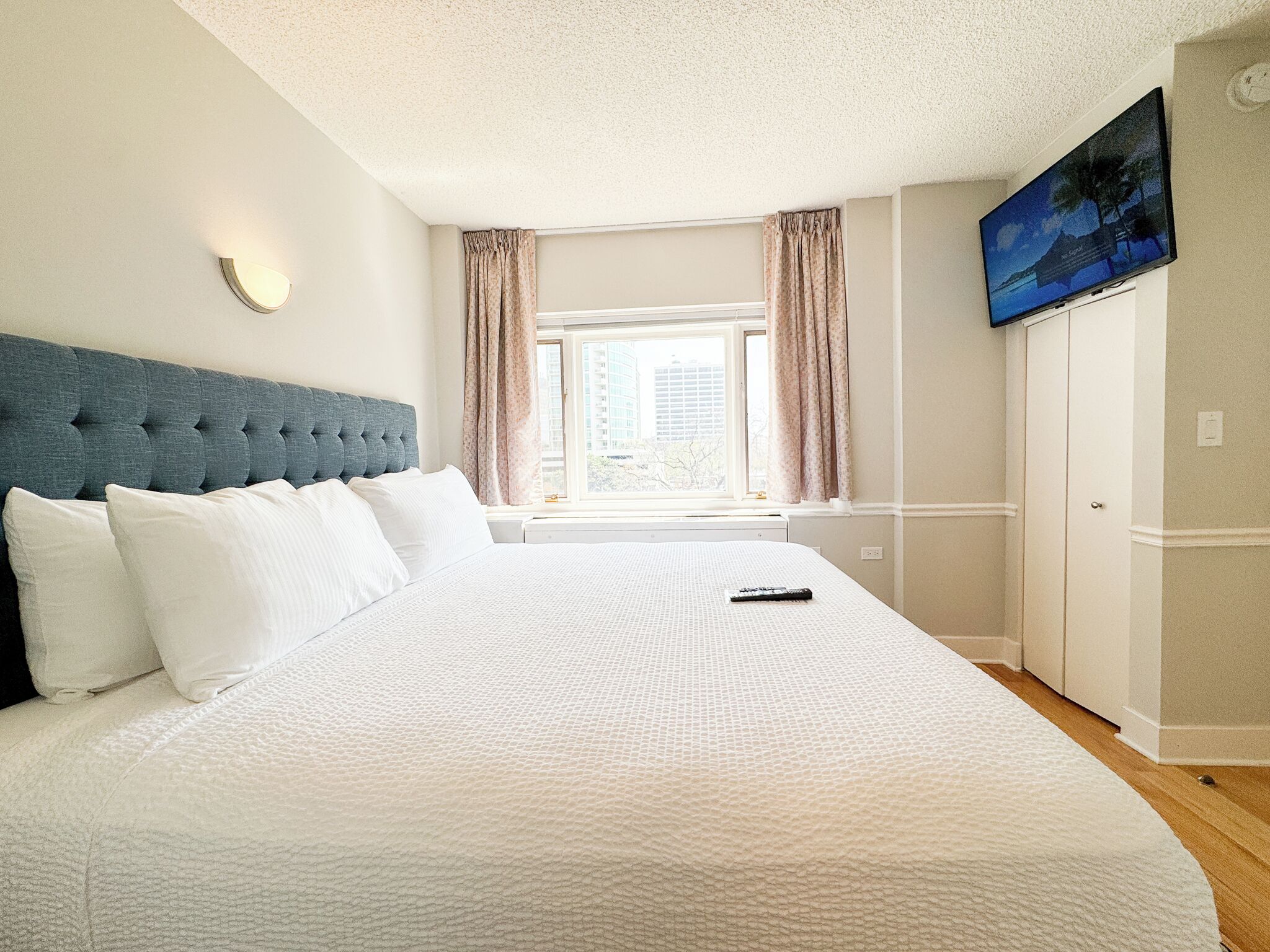 Un dormitorio con una cama grande, cabecera, ventana con cortinas, televisor de pantalla plana en la pared y vista a la ciudad.