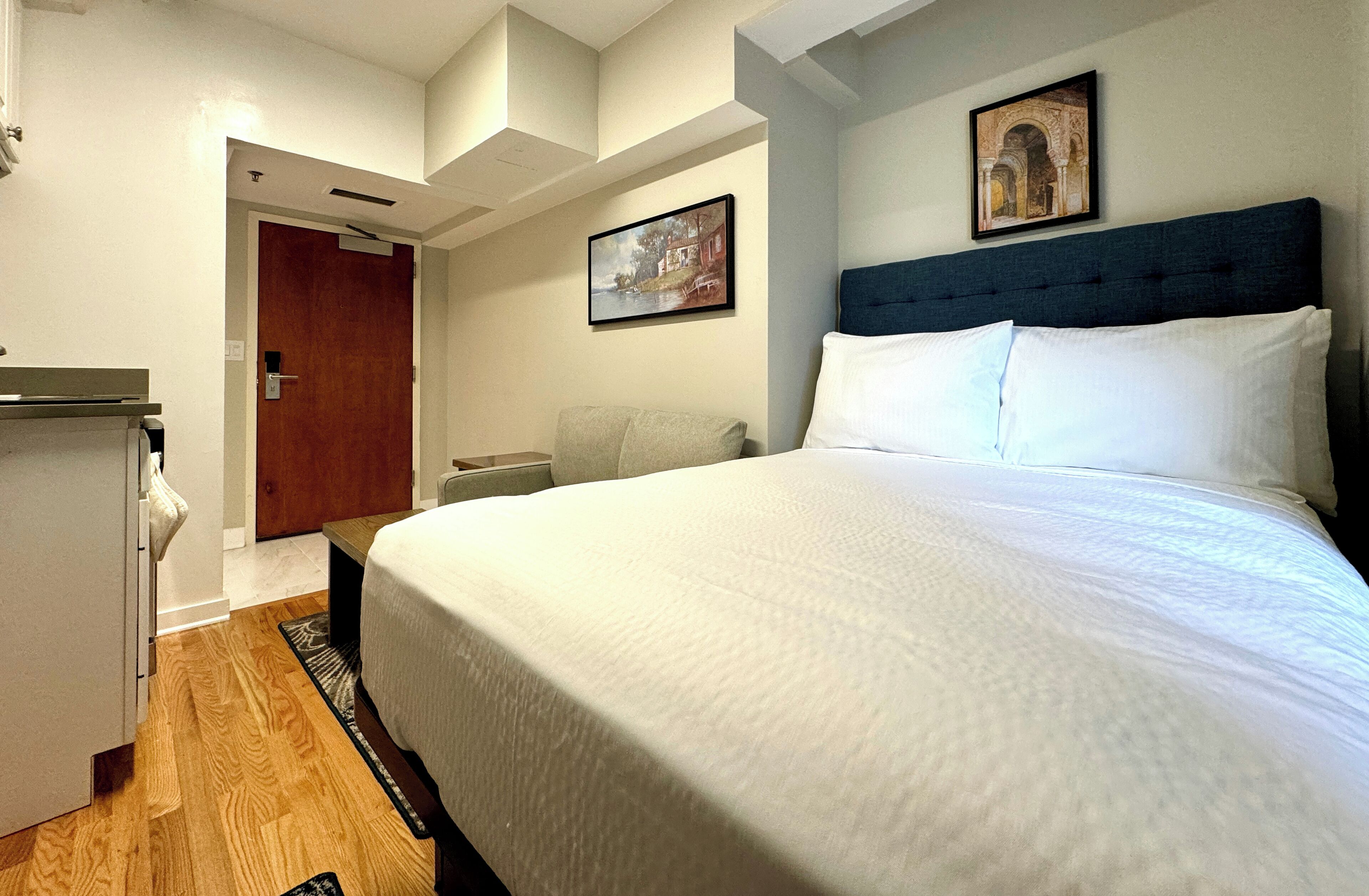 Habitación de hotel con una cama grande, un escritorio y una silla. Se ve una puerta que da a otra habitación y un cuadro en la pared.