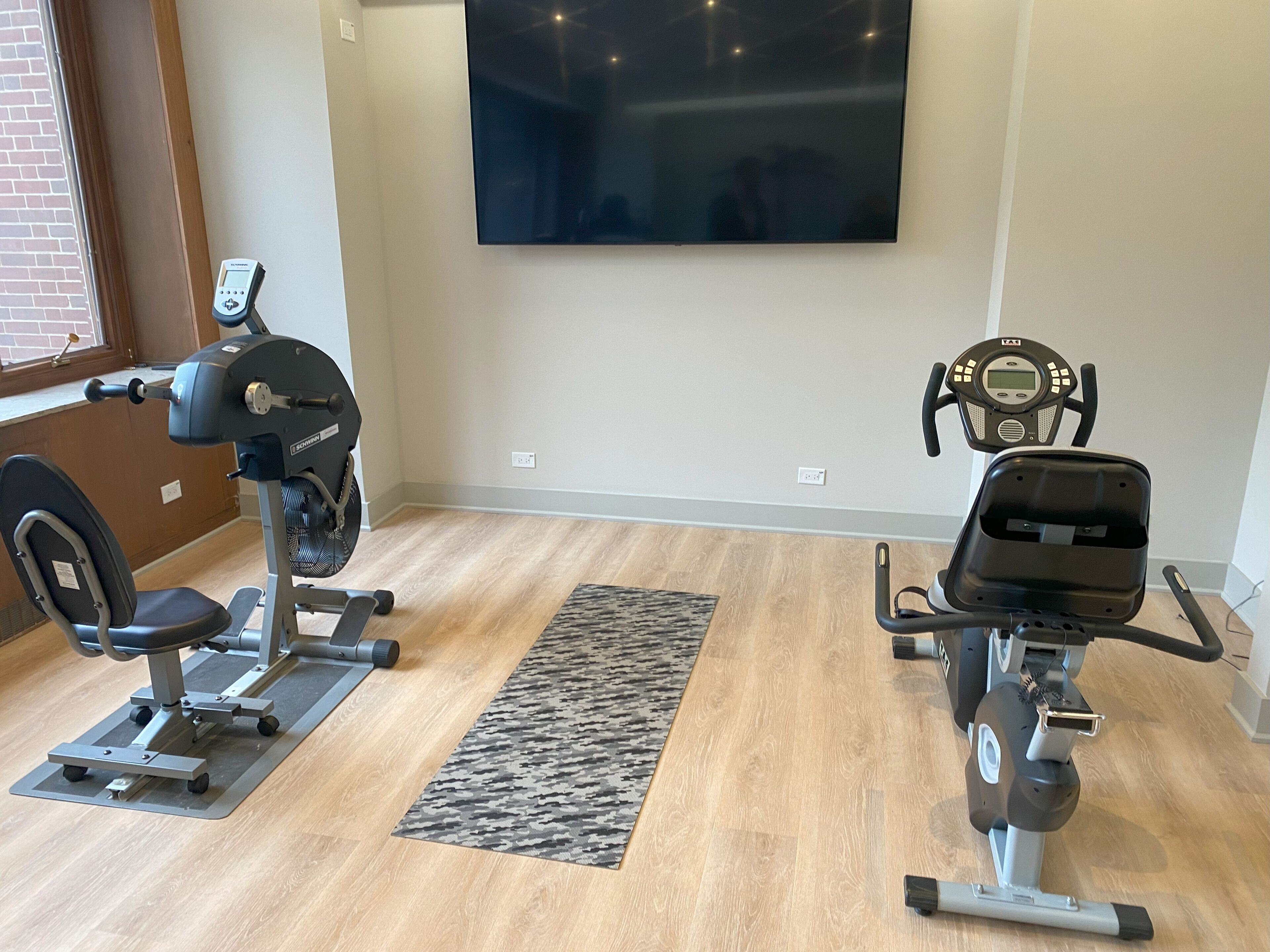 A home gym with two exercise bikes, a TV mounted on the wall, and a patterned rug on the floor.
