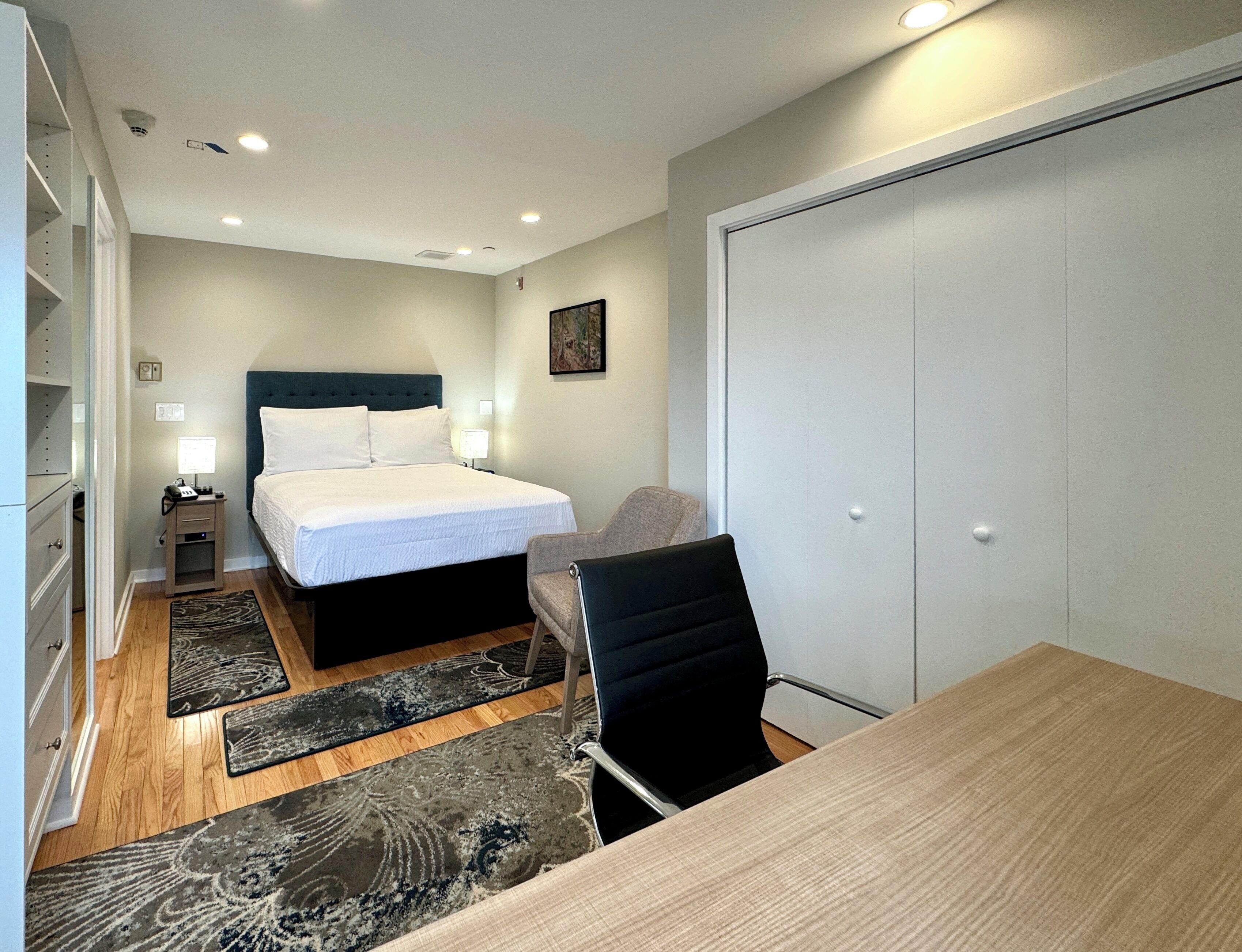 A hotel room with a bed, bedside tables, a desk, a chair, a wardrobe, and a carpeted floor.