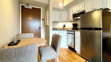 Una cocina funcional con armarios blancos, un refrigerador de acero inoxidable y una zona de comedor con mesa de madera y dos sillas grises.