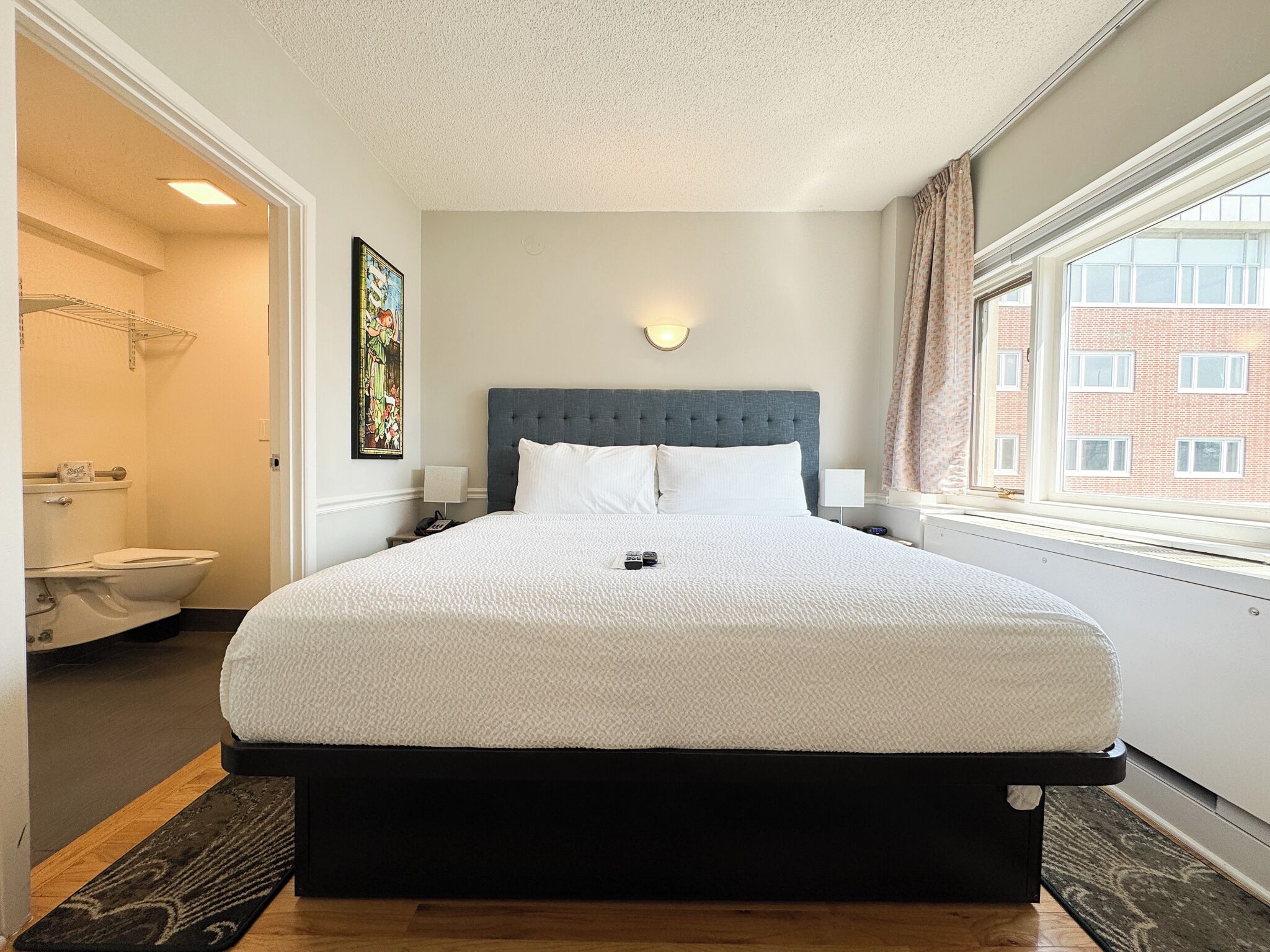 A bedroom with a large bed, a headboard, a bedside table, a nightstand, a window with curtains, and a view of a building outside.