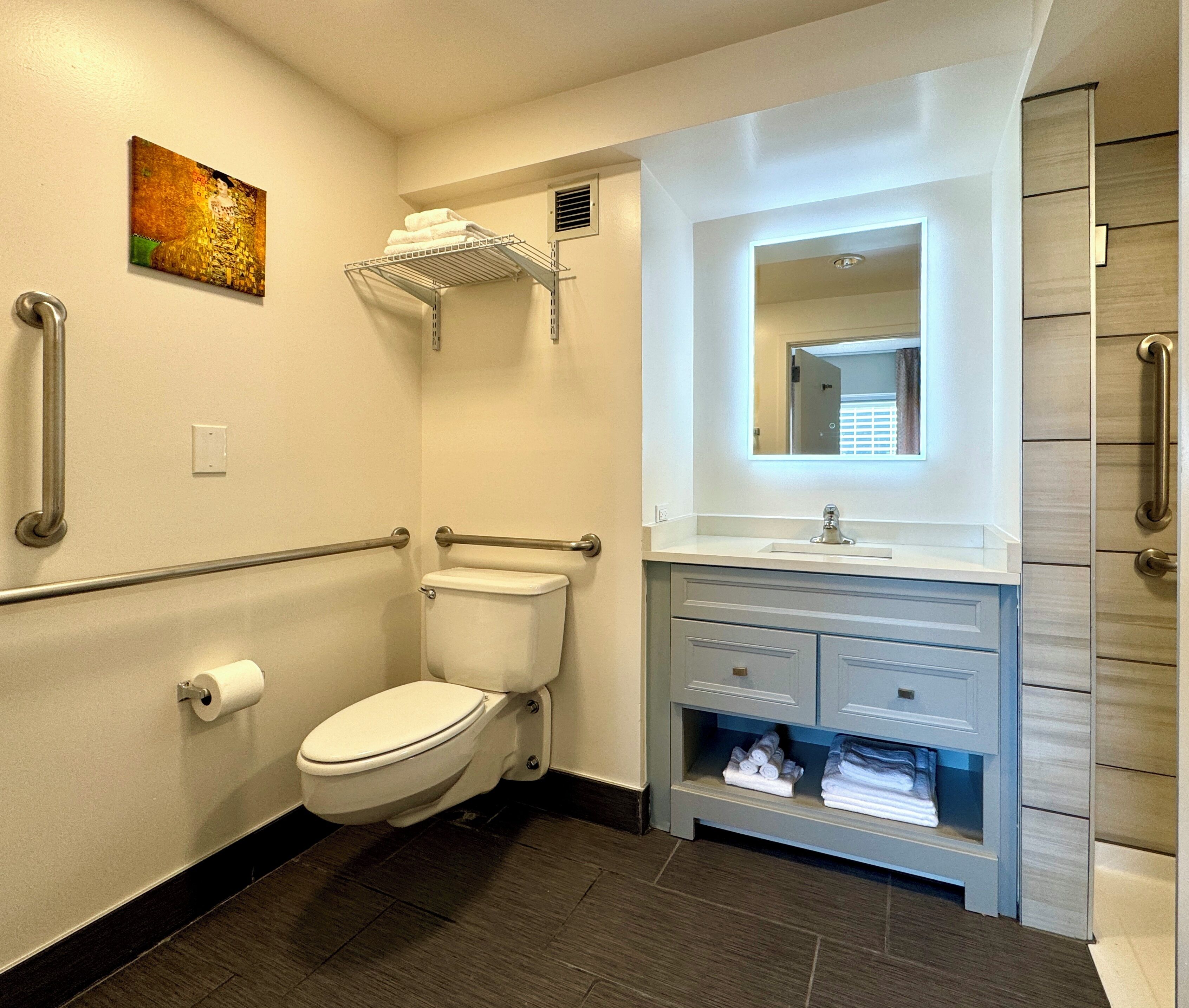 A modern bathroom with a toilet, sink, and vanity. There are grab bars on the walls and a towel rack above the toilet.