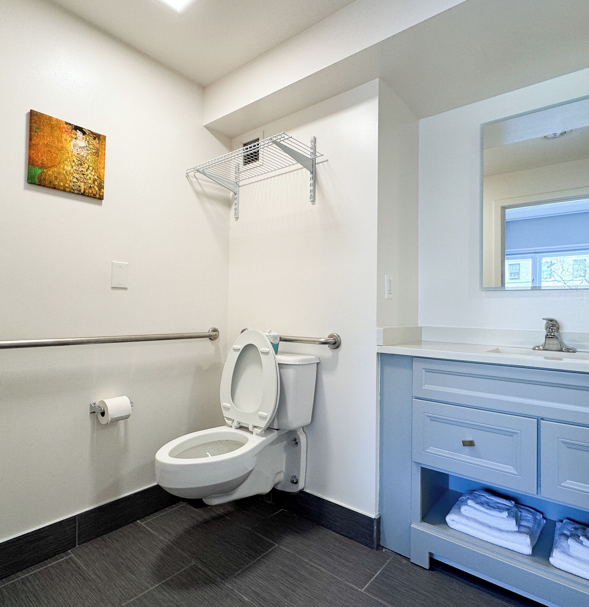 Et moderne badeværelse med toilet, metalhylde over toilettet, hvid vask med spejl og et håndklædetørrestativ.