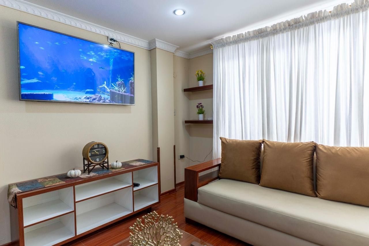A living room with a flat-screen TV mounted on the wall, a sofa, a wooden console table, and a decorative plant.
