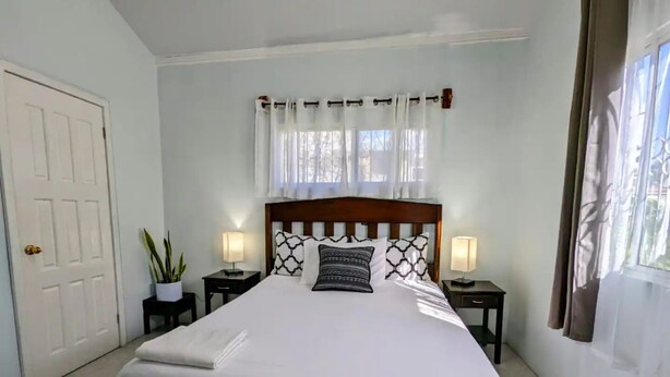 A neatly arranged bedroom with a large bed, two bedside tables with lamps, a window with curtains, and a door. - Elle's 2 BR/2 bathroom, home in San Ignacio. Minutes from town & Maya Ruins (Cayo District)