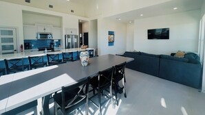 A modern kitchen with white cabinetry, a dining area with a long table and chairs, and a living area with a sofa and a flat-screen TV. - Gated 5 Bed • 4 Bath • Private Beach Access  • Early Check-In & Late Checkout (Bolivar Peninsula)
