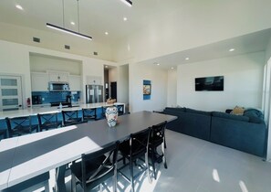 A modern kitchen with white cabinetry, a dining area with a long table and chairs, and a living area with a sofa and a flat-screen TV.