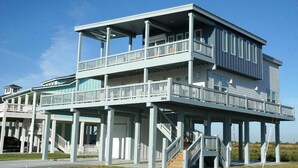 A two-story beachfront house with a balcony on the upper floor, a covered porch on the ground floor, and a staircase leading to the upper level. - Gated 5 Bed • 4 Bath • Private Beach Access • Early Check-In & Late Checkout (Bolivar Peninsula)