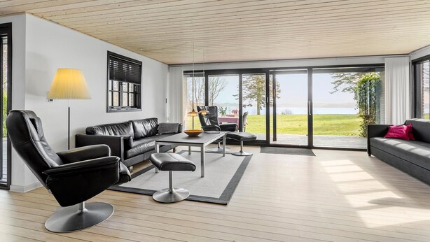A modern living room with a sofa, chairs, a coffee table, and a lamp. There is a large sliding glass door leading to an outdoor area with a view of a body of water. - Awesome home in Roslev with kitchen (Roslev)