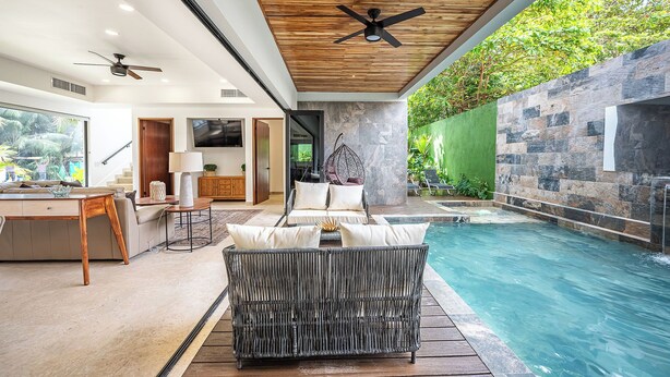 A modern indoor pool area with a wicker sofa, a wooden ceiling fan, and a stone wall. - Brand New Luxury Villa on the South End of Jaco Beach, Steps from the Beach! (Jaco)
