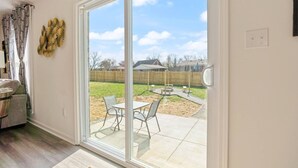 Ein Zimmer mit einer Schiebetür zu einer Terrasse im Freien mit Tisch und Stühlen, einem Holzzaun und einem wolkenlosen Himmel.