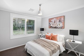 A bedroom with a bed, two bedside tables, a window with blinds, a ceiling light, and a wall-mounted painting.