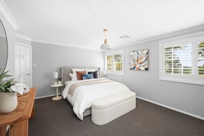 A bedroom with a bed, bedside table, lamp, artwork, window, and a plant.