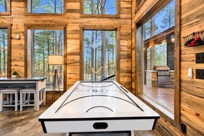 A wooden cabin interior with a central table, bar seating, a lamp, and a view of the forest through large windows.