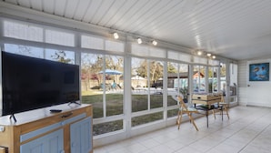 A spacious room with large windows, a wooden cabinet with a flat-screen TV, and a children's play table. - Mermaid Dreams: Spacious 3 bedrooms 2.5 bath with pool ALL in golf cart district (Ocean Springs)